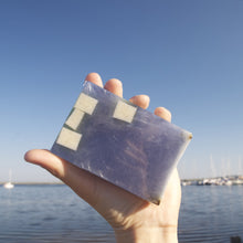 Charger l'image dans la galerie, Le savon Exfolient Doux est pris en photo devant un paysage maritime, avec la petite baie et des bateaux à voile en arrière plan.