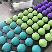Charger l'image dans la galerie, Plusieurs bombes de bain fraîchement pressées sont sur la table de production. Il y a un lot de couleur turquoise, un lot vert vif et un lot mauve améthyste.