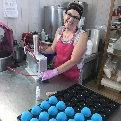 Mylène, notre assistante à la production, sourit tout en pressant des bombes de bain. Elle utilise une presse pneumatique et porte un tablier rose. Les bombes de bain sont d'un beau bleu ciel.