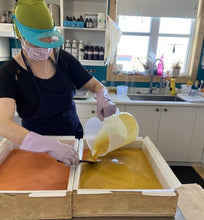Charger l'image dans la galerie, Ariane, notre savonnière, est entrain de couler la couche jaune du savon arc-en-ciel dans un grand moule en bois chemisé.