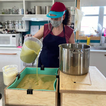 Charger l'image dans la galerie, Ariane, notre savonnière, est entrain de faire le motif marbré du savon avocat crémeux. Elle tient un pichet de savon frais qu'elle verse dans le grand moule à savon.