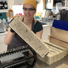 Charger l'image dans la galerie, Ariane, notre savonnière, tient une longue brique de savon Noir Hybride SAfrcain, juste avant la coupe. Sur la table de travail, on voit la tanche à savon et d'autres longues briques de savons.