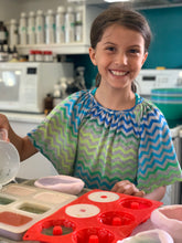 Charger l&#39;image dans la galerie, Une jeune fille sourit pendant qu&#39;elle participe à un atelier de fabrication de savon. On voit des moules et du savon frais devant elle sur la table de travail.
